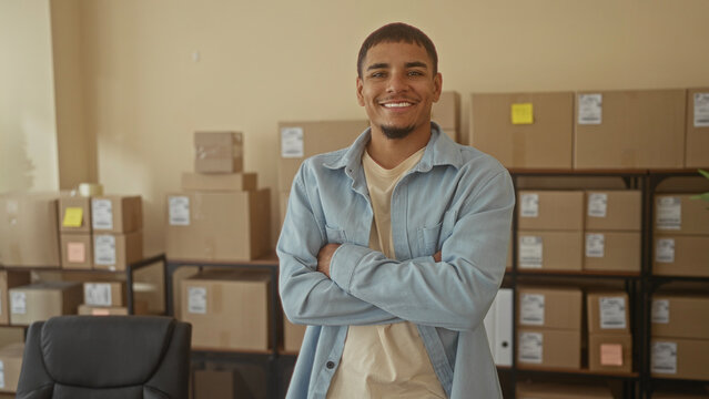 Young man with arms crossed smiling in a building surrounded by stacked cardboard boxes and shipping labels; small business success pride.