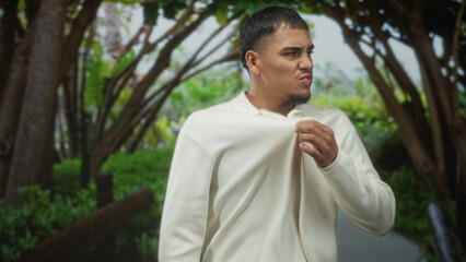 Man in cream sweater with hand on chest, adjusting collar and walking while looking sideways on a forest path; calm reflection.