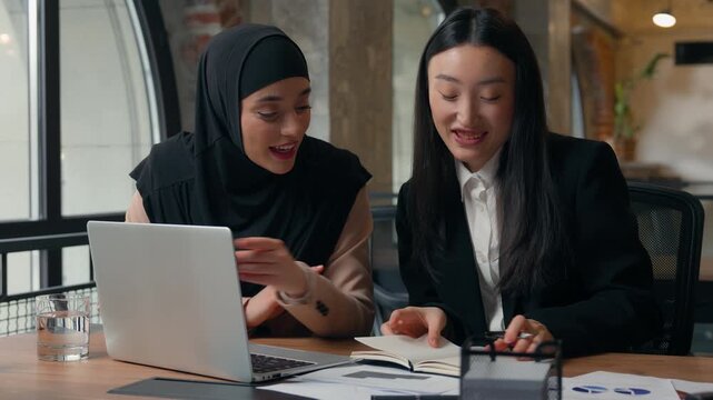 Happy smiling carefree two multiracial girls team business women diversity multiethnic businesswomen female in office working online together Muslim leader help Asian intern with laptop writing notes