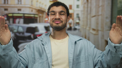 Young hispanic man in glasses raises both hands with palms outward on sunlit busy city street;...