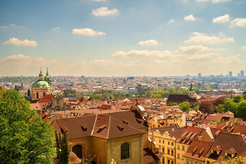 Panoramic view of Prague skyline with red tiled roofs, St. Nicholas Church green dome, and modern city skyline in distance under blue sky © Arpan