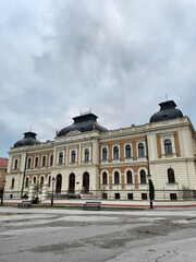 Obraz premium Elegant architectural facade of a historic building under cloudy sky, viewed from an open plaza with benches and soft natural light. Sremski Karlovci, Serbia.