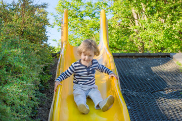 Happy cute little child boy having fun on a yellow slide outdoor in the park, sunny summer day in children playground. Selective focus
