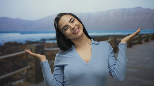 Young woman smiling playfully with hands covering and uncovering her face on a seaside promenade under blue skies by the ocean - Powered by Adobe