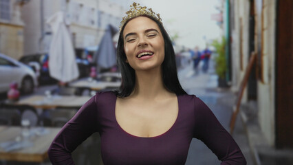 Young woman with crown smiling on outdoor terrace in street setting near restaurant and coffee shop.
