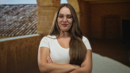 Naklejka premium Woman with arms crossed in old town building, wearing glasses and white tee; confidence independence self reliance.