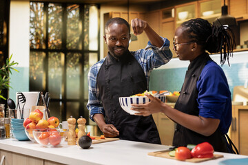 Cheerful couple adding pinch of salt to the salad in the kitchen, preparing food with fresh organic ingredients. Man being funny imitating the chef from social media trends, joy and wellness.