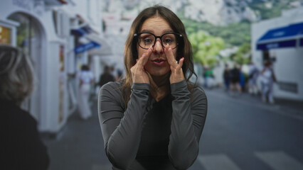Woman wearing glasses cupping hands around mouth shouting on street sidewalk by storefronts and a crosswalk while leaning forward; urgency alert.