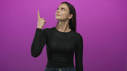 Woman points index finger upward in magenta studio while looking up and wearing black shirt and jeans; curiosity.