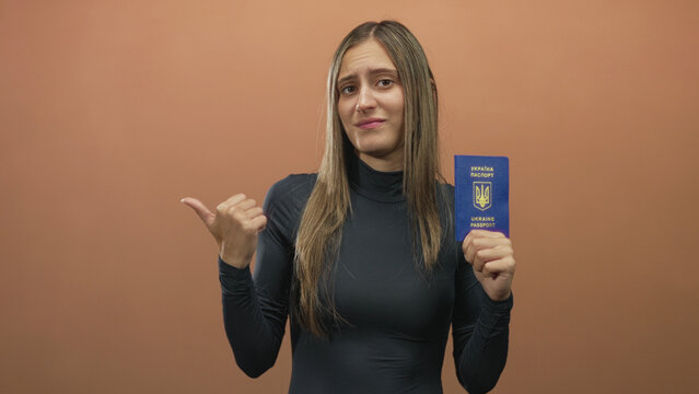 Young hispanic woman holding ukraine passport and pointing thumb to side in studio; identity travel skepticism.