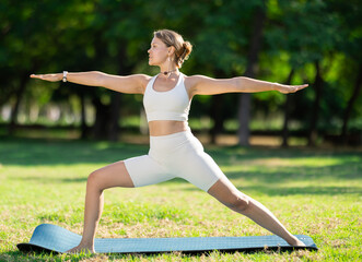 Fototapeta premium Motivated young girl performing standing lunging asana Virabhadrasana, or Warrior pose during outdoor yoga session on green glade in summer park