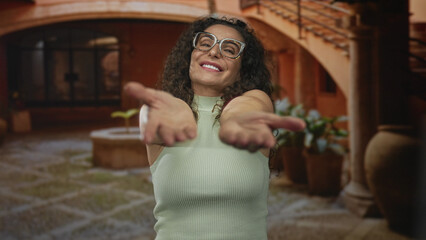Woman extends hands toward camera smiling in building courtyard under archway; open friendly welcome invitation.