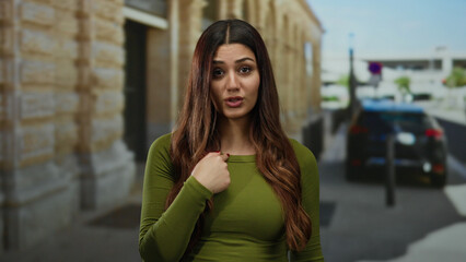Fototapeta premium Woman outdoors gestures no with finger in urban street scene, exuding calm confidence, wearing green, hispanic descent, showcasing casual fashion against blurred city background.