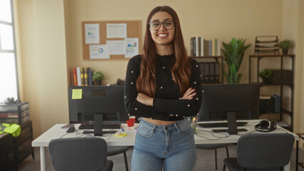 Hispanic woman with crossed arms standing confidently in office room with computers and bookshelves, showcasing professional indoor workplace environment.