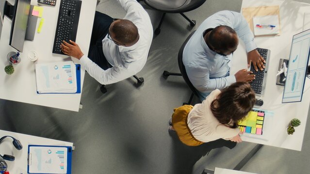 Top down view of business people review diagrams during report writing. Colleagues in multinational company share insights and coordinate tasks using pc and visual data tools.