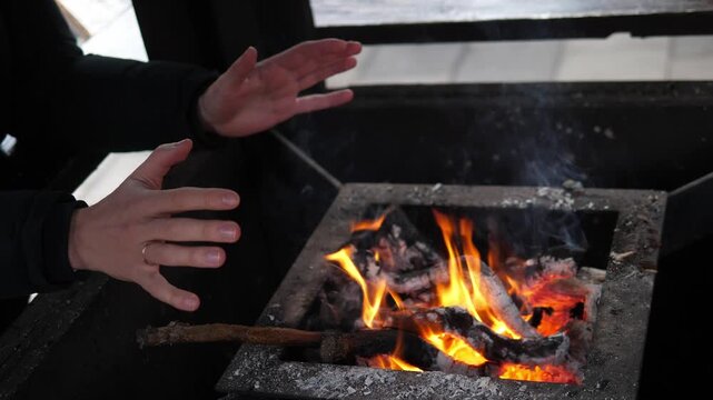 A man warming their hands over a small open fire at a Christmas market. The glowing flames create a cozy contrast to the cold winter atmosphere surrounding the festive outdoor event