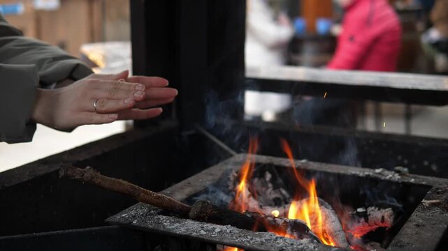 A woman warming their hands over a small open fire at a Christmas market. The glowing flames create a cozy contrast to the cold winter atmosphere surrounding the festive outdoor event