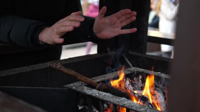 A man warming their hands over a small open fire at a Christmas market. The glowing flames create a cozy contrast to the cold winter atmosphere surrounding the festive outdoor event