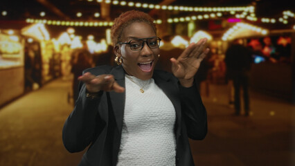 Woman wearing glasses and gold hoop earrings smiling and finger pointing to colorful souvenir stall on busy city street market at night; joy.