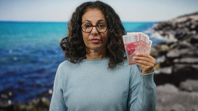 Middle-aged woman with icelandic krona in hand standing on a seaside landscape, highlighting economic themes against a serene beach backdrop.