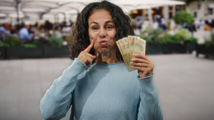 Middle-aged woman at outdoor cafe terrace playfully puffing cheeks holding brazilian banknotes creating a joyful scene with urban background and diverse people around.