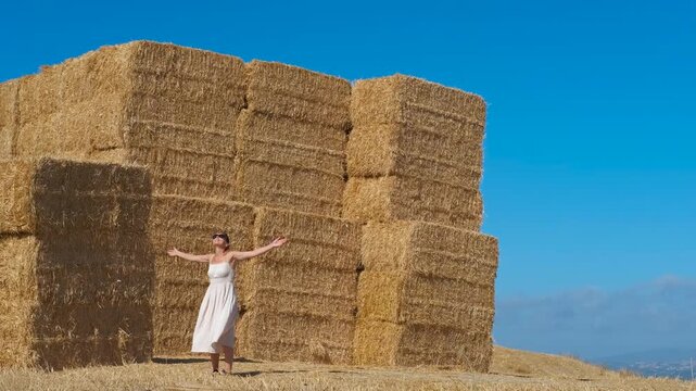 Free woman spinning happily near hay bales in summer. Young woman in a white dress spinning with her arms outstretched, feeling free and joyful near a large stack of hay bales against a clear blue sky