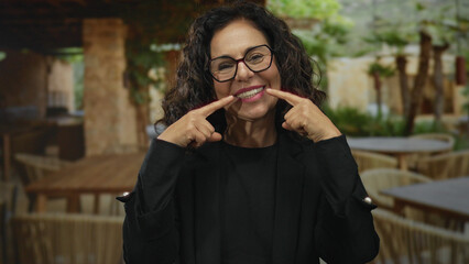 Woman smiling at a terrace coffee shop, wearing glasses and black outfit, pointing at her smile, with blurred outdoor seating in the background, creating a cheerful atmosphere.