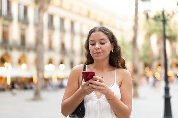 Obraz premium Young smiling woman with smartphone walks along Royal square, walks around city, spend time in public urban space. Electric street lighting, twilight in southern city, evening life in big city ..