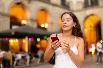 Obraz premium Young smiling woman with smartphone walks along Royal square, walks around city, spend time in public urban space. Electric street lighting, twilight in southern city, evening life in big city ..