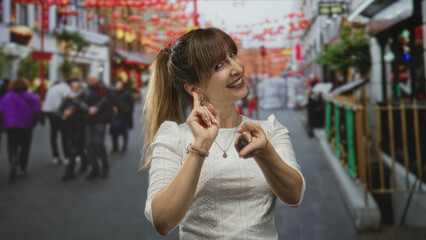 Woman points finger to camera on a busy city street market while smiling and showing hand gesture; joyful invitation.