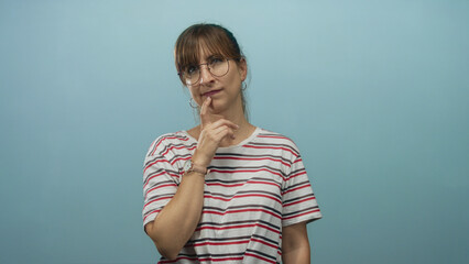 Woman wearing striped shirt and glasses with finger to lips for silence in studio with light blue wall; curiosity pondering.