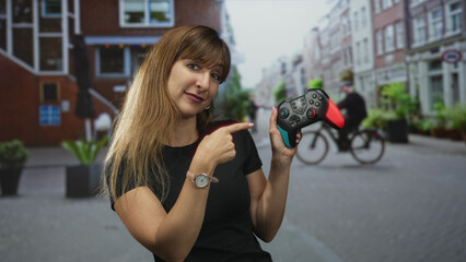 Woman holding game controller and points index finger in city street, smiling with relaxed pose and visible nails; playful game demo.