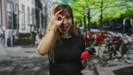 Woman holding red gamepad and making ok sign to eye on city street with parked bicycles and cars in view  curiosity. © Krakenimages.com