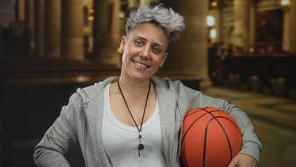 Caucasian woman holding a basketball while leaning against a pew inside church building with whistle around neck; confidence.