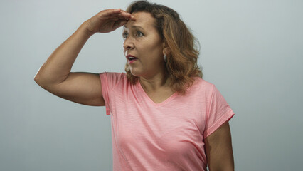 Middle age hispanic woman shielding forehead with hand in a gray studio setting to shade eyes while scanning for distant views; curiosity discovery exploration wonder.