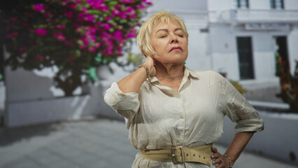 Fototapeta premium Senior hispanic woman touching neck for neckache on a sunlit white street by pink bougainvillea; discomfort rest.
