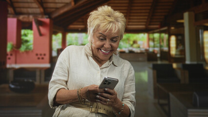 Woman holding smartphone and tapping screen while seated in building reception area, smiling and reading messages; digital connection contentment.