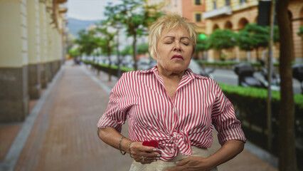 Senior hispanic woman holding small red heart near chest, eyes closed, on street lined with trees and buildings; heartache.