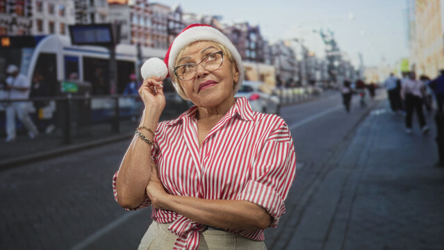 Woman in santa hat with hand on chin thinking on a busy city street by a tram and cars, arms crossed and wearing a red striped shirt; pensive reflection. - Powered by Adobe