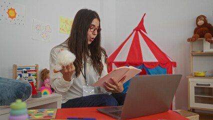 Hispanic woman teacher reading to preschool children in a vibrant kindergarten setting with toys tent and laptop
