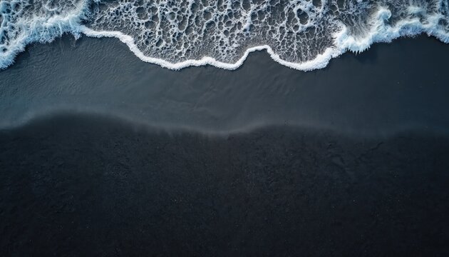 Top view of ocean wave washing ashore on dark black sand beach. White sea foam creates intricate patterns on the wet coastline. Natural beauty abstract background.