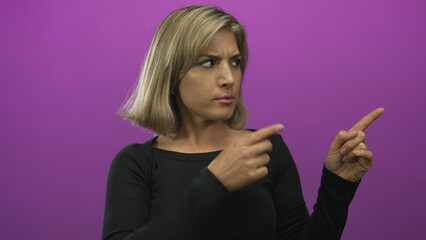 Young blonde woman posing casually pointing finger to the right in purple studio under soft...