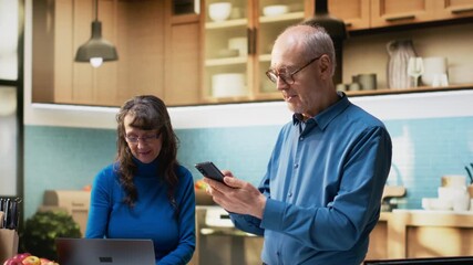 Senior couple checking smartphone apps in home kitchen with a relaxed smile, enjoying casual conversation and laughing with their phone. Peaceful retirement lifestyle together. Camera B.