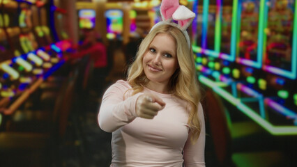 Fototapeta premium Blonde woman wearing bunny ears pointing while smiling in vibrant casino setting with colorful slot machines creating a playful and engaging atmosphere.