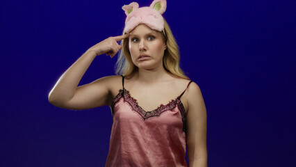 Woman in nightwear making a crazy gesture with her finger to her head, wearing a pink sleep mask against a blue background, conveying confusion or playfulness.