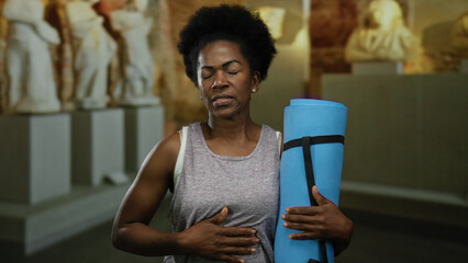 Woman feeling discomfort holding yoga mat in museum surrounded by sculptures suggesting an indoor setting of tranquility and introspection.