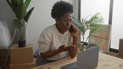 Woman talking on phone in living room with laptop amid moving boxes and plants, embodying a mix of communication and the excitement of settling into a new apartment.