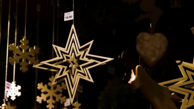 Handcrafted wooden star and angel decorations with price labels hanging and swinging in warm festive lighting at a christmas market stall.
