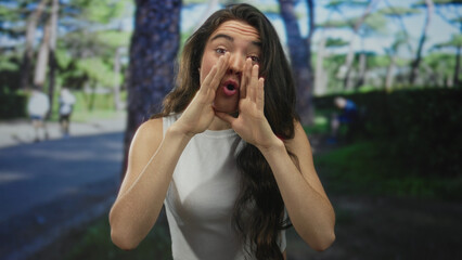 Naklejka premium Young hispanic woman cupping hands around mouth shouting amid green forest under sunlight; enthusiasm.