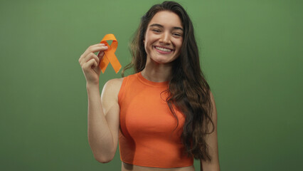 Smiling young woman wearing orange top holds orange ribbon in green studio; awareness support hope solidarity.